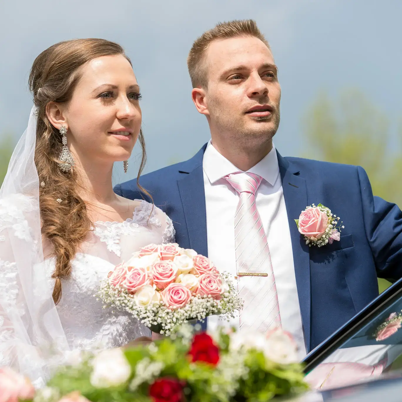 Ein Brautpaar steht draußen an ihrem Hochzeitsauto bei blauem Himmel